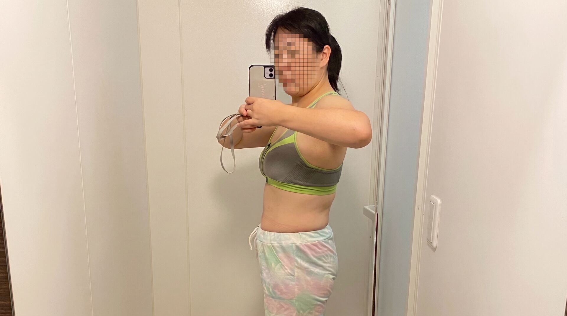 19kgの大幅な減量に成功し、体重71kgを達成した40代女性の全身を横から写した鏡越しのセルフィー。彼女はグレーにネオンイエローのラインが入ったスポーツブラと、パステルカラーのマーブル柄ショートパンツを着用し、髪をポニーテールにまとめています。両手でスマートフォンを持ち、真剣な表情で画面を見つめています。すっきりと引き締まったお腹周りや姿勢から、これまでのダイエットの努力と見事な成果がはっきりと伝わってくる1枚です。背景はシンプルな白い壁とタイルの床です。